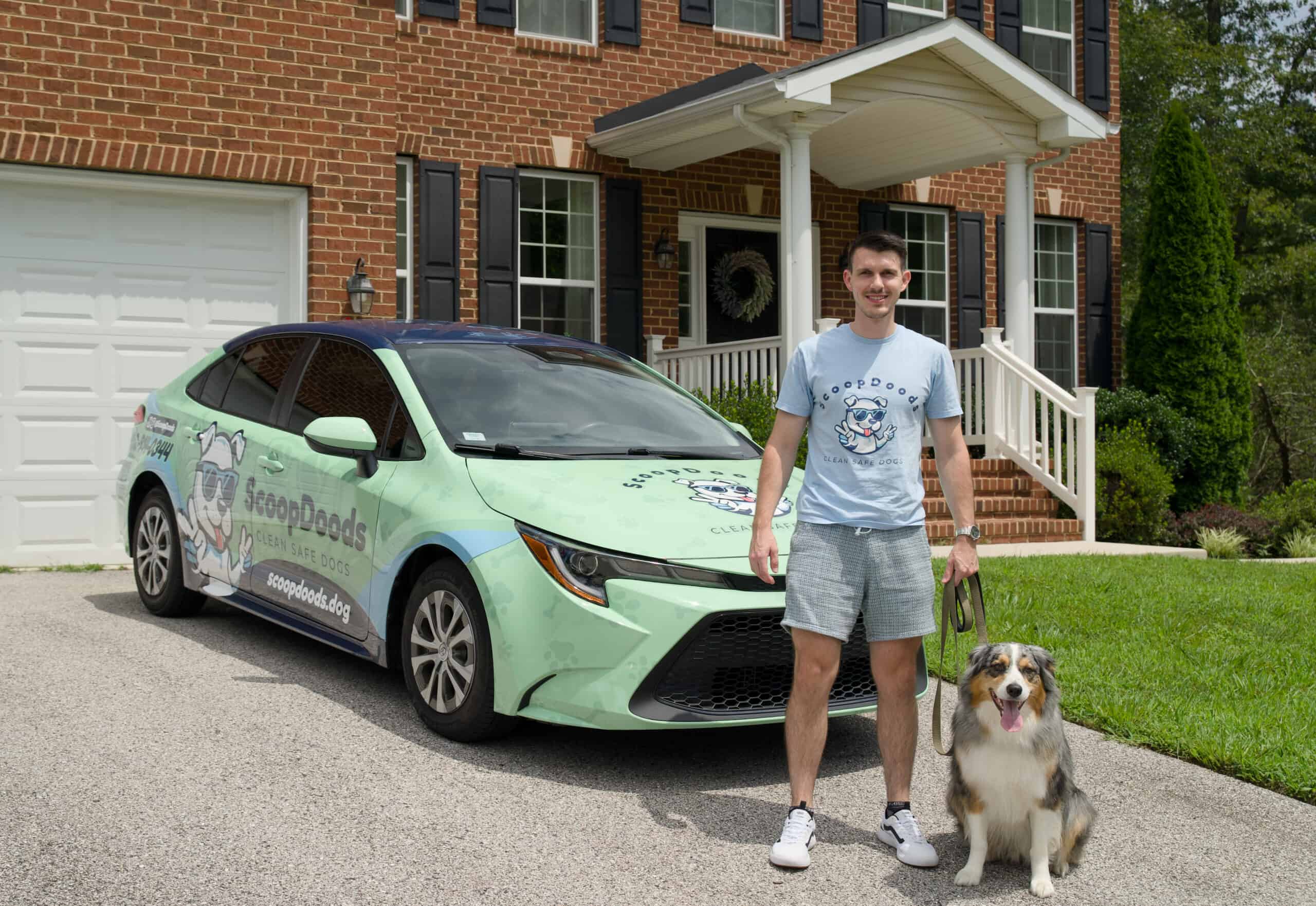A man in a light blue ScoopDoods t-shirt stands in front of a ScoopDoods-branded car while holding the leash of a happy dog outside a brick house.