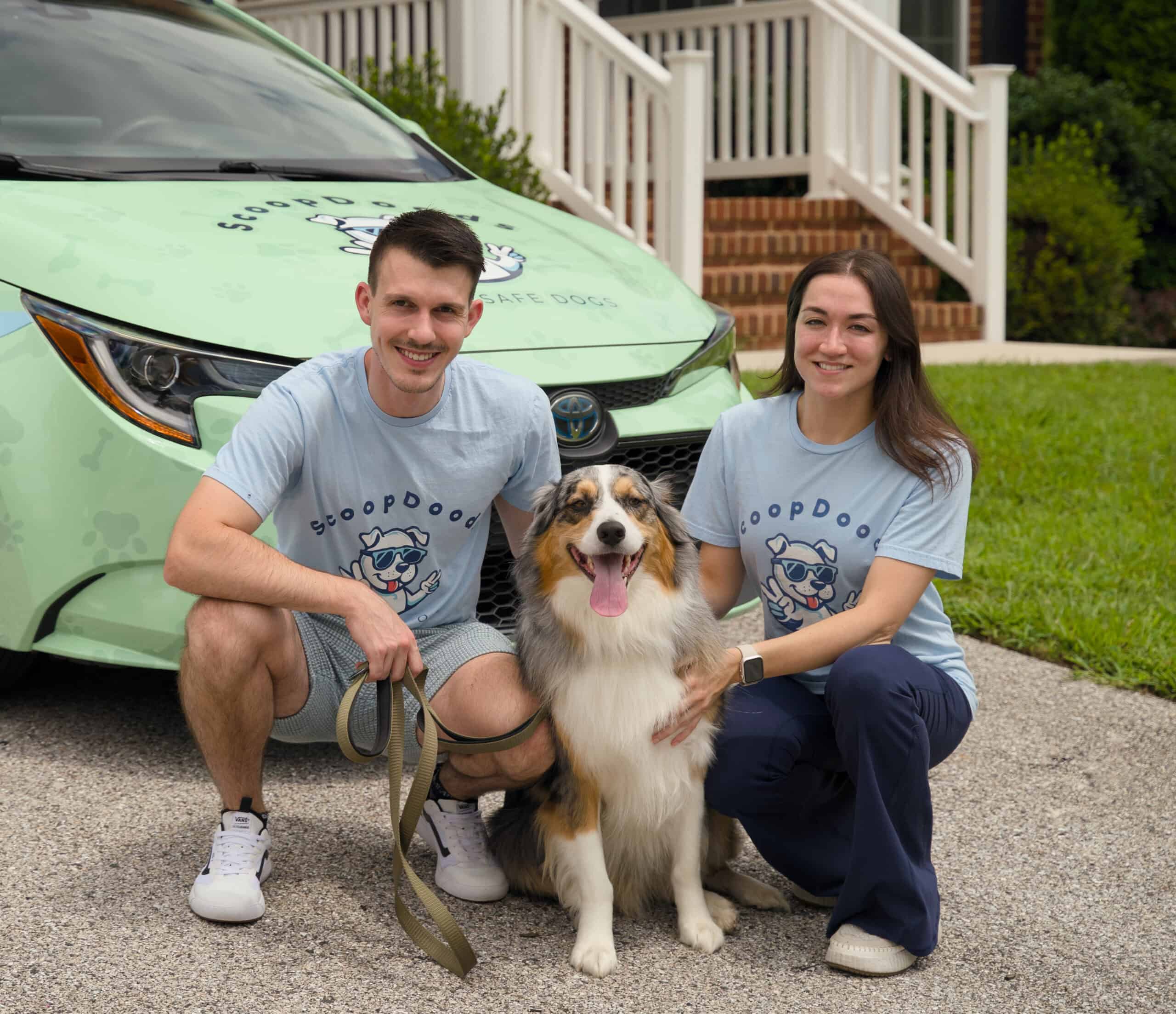 A man and woman in matching ScoopDoods t-shirts kneel beside a happy dog in front of a branded ScoopDoods car.
