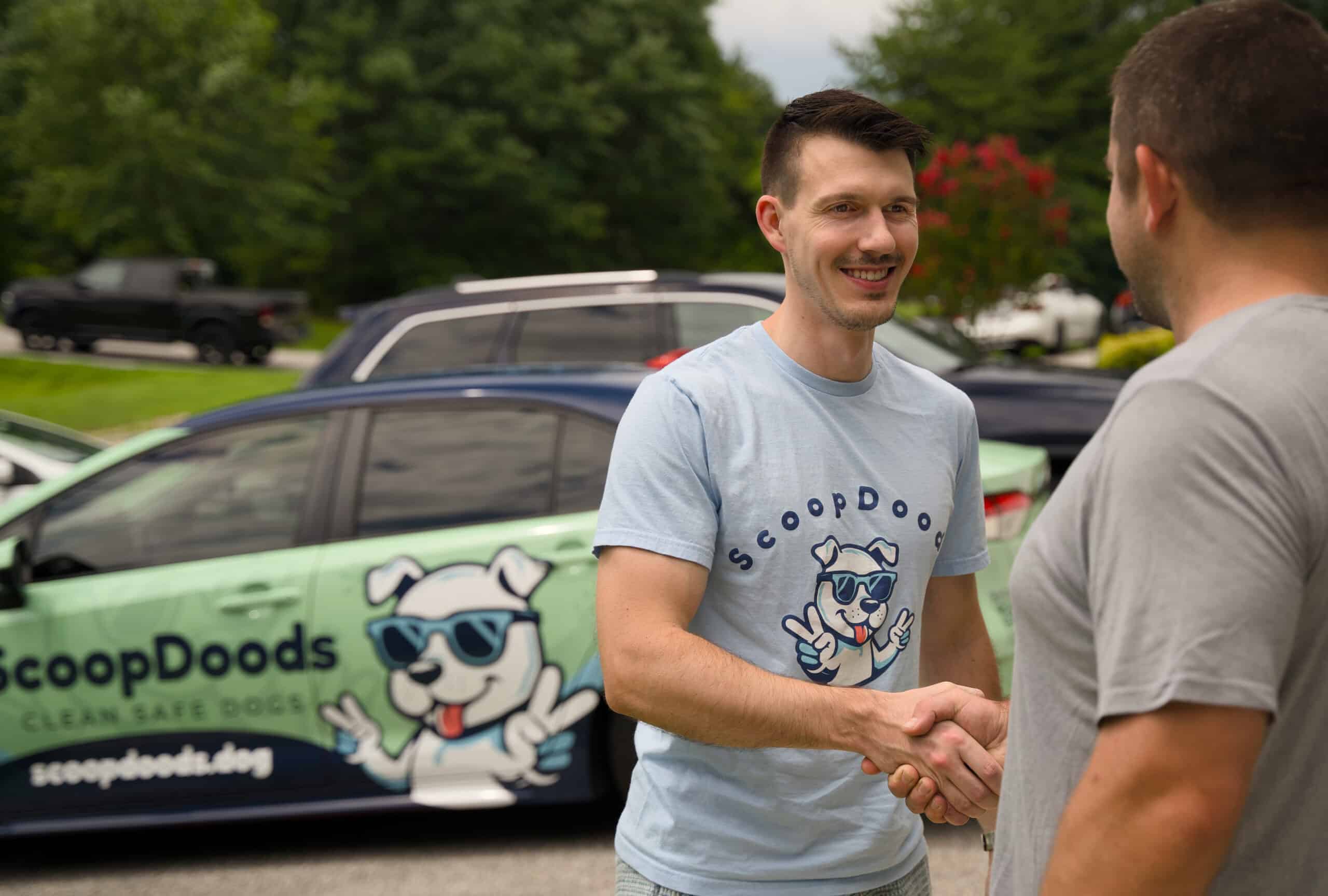 A man wearing a light blue “ScoopDoods” t-shirt smiles while shaking hands with another person in front of a car wrapped with the ScoopDoods dog mascot and branding.
