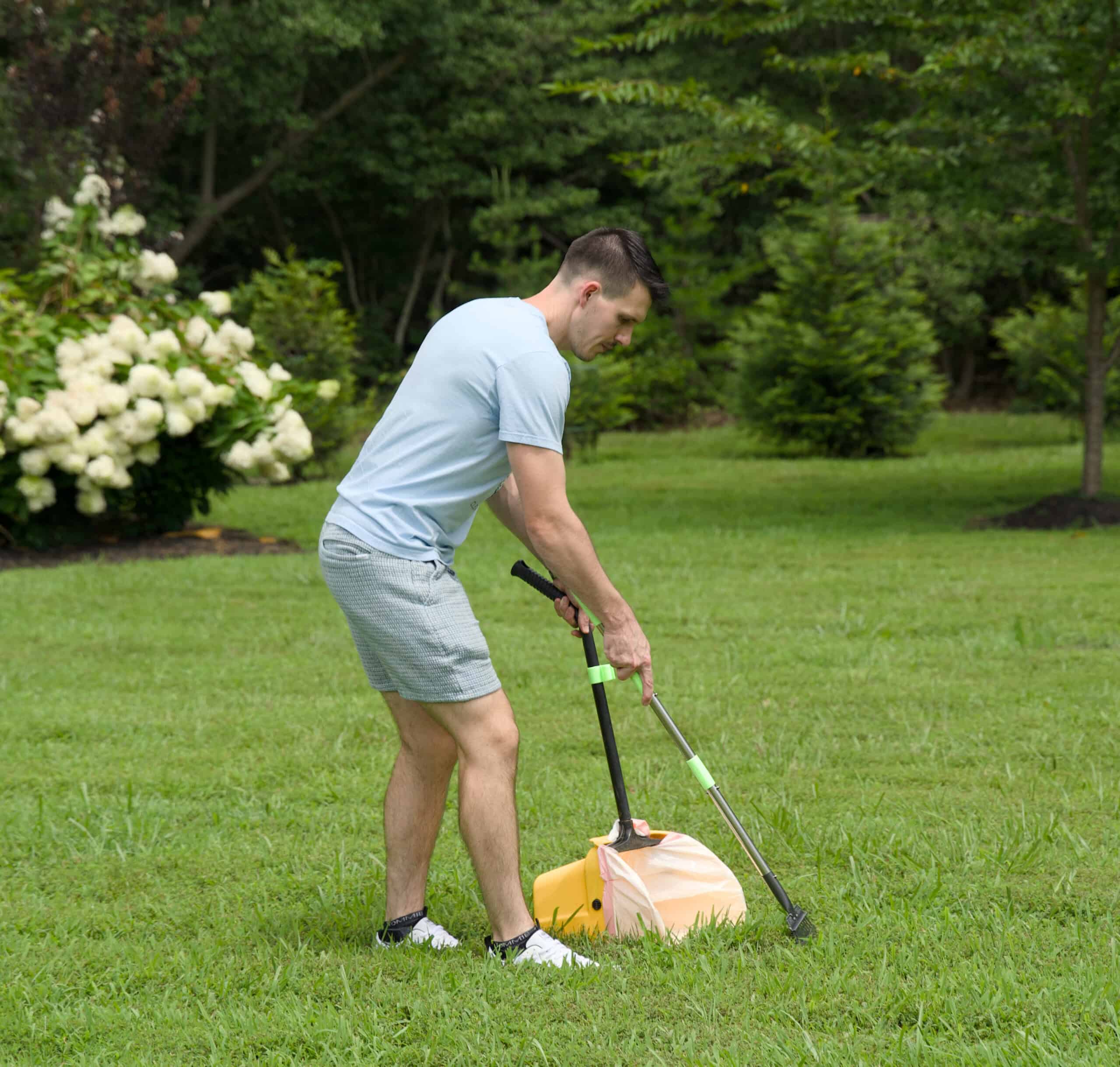 A man in a light blue t-shirt and shorts uses a pooper scooper tool to clean up a yard, collecting waste into a lined bin.